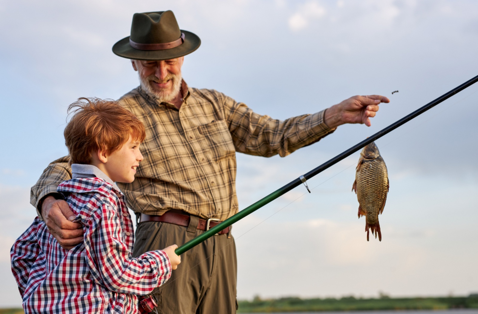A senior holds a caught fish on the end of a fishing line as their young grandchild holds the fishing rod and smiles
