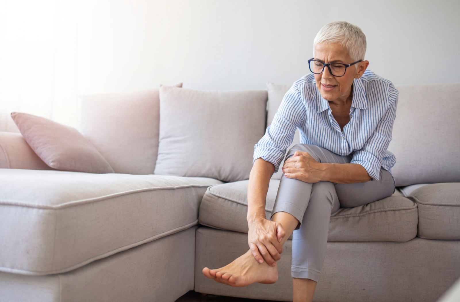 A senior sits on a sofa and rubs their ankle to try and reduce swelling