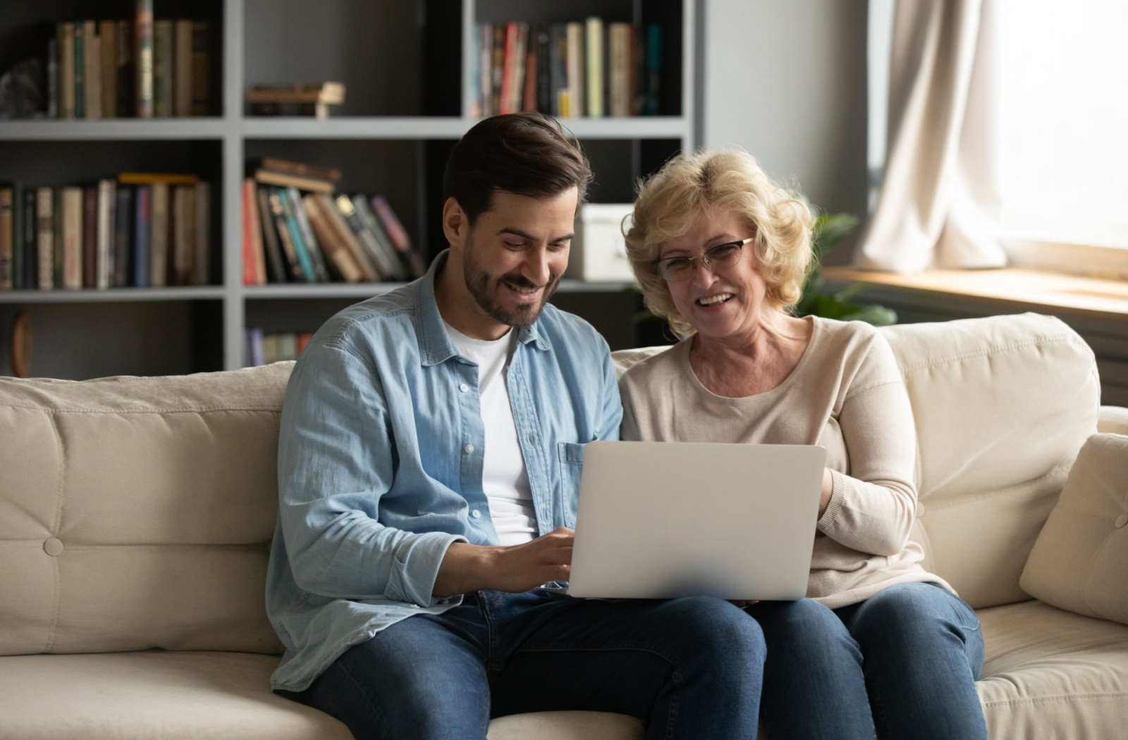 An adult child and their senior mother smile as they browse senior living options on a laptop while sitting on a sofa together.
