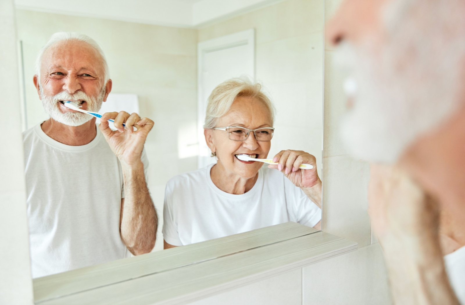 A senior couple brushes their teeth together while looking in the mirror 