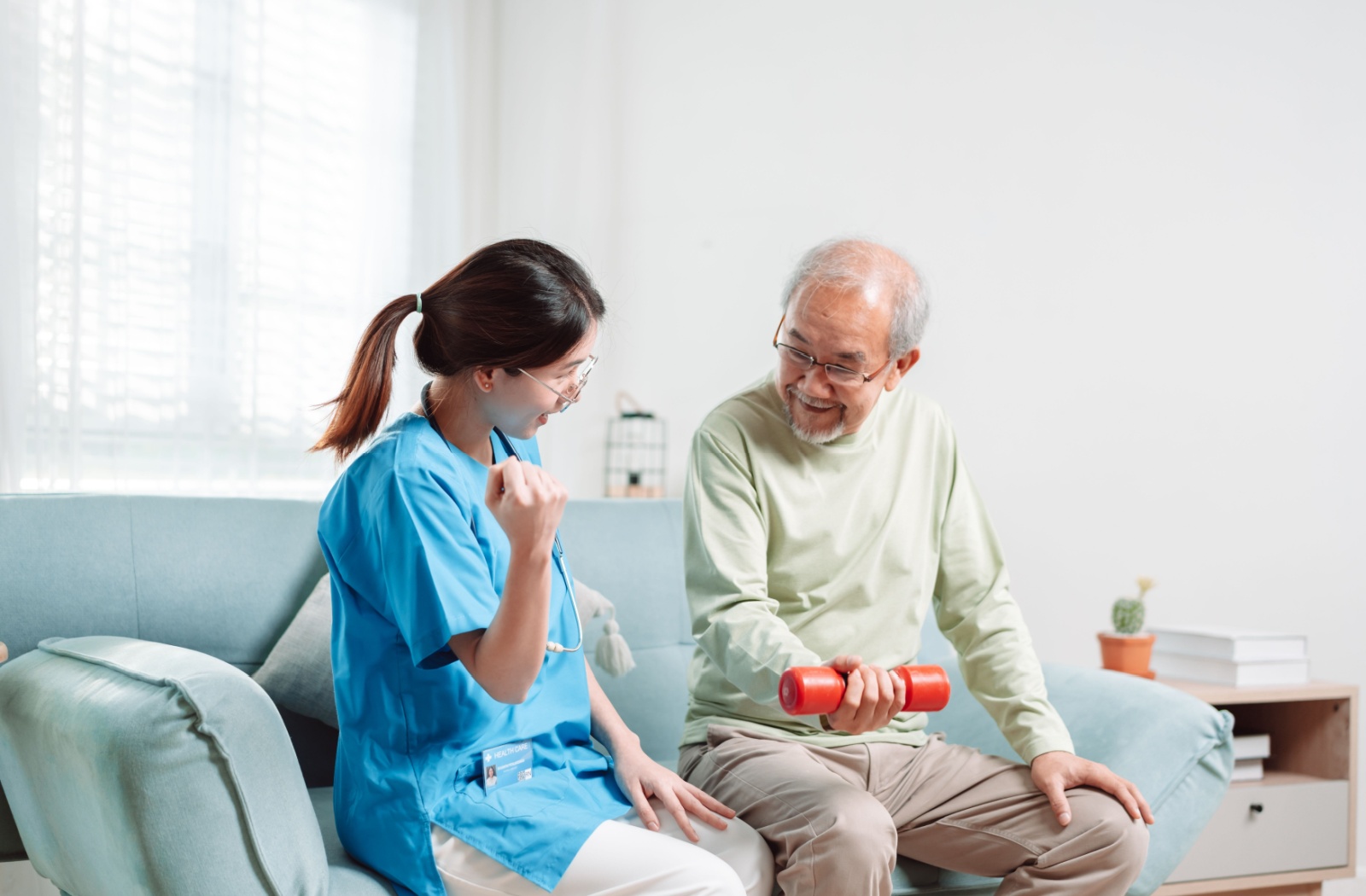 An assisted living caregiver helps a senior resident with low-impact arm exercises using a red dumbbell