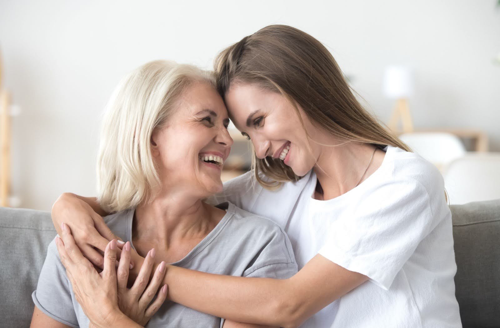 An adult child comforts their exasperated senior parent while they sit on a tan sofa.