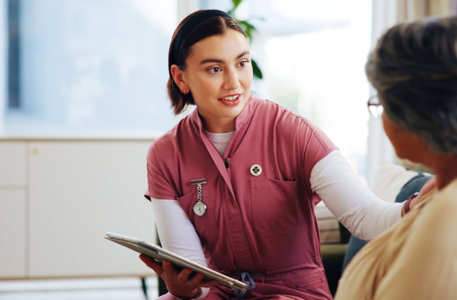 A caregiver wearing berry-colored scrubs and holding a clipboard comforts a senior resident.