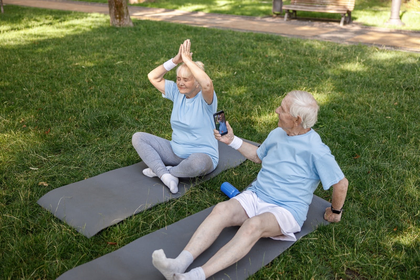 Senior older man taking a picture of his wife smiling and doing yoga asana on field grass in park