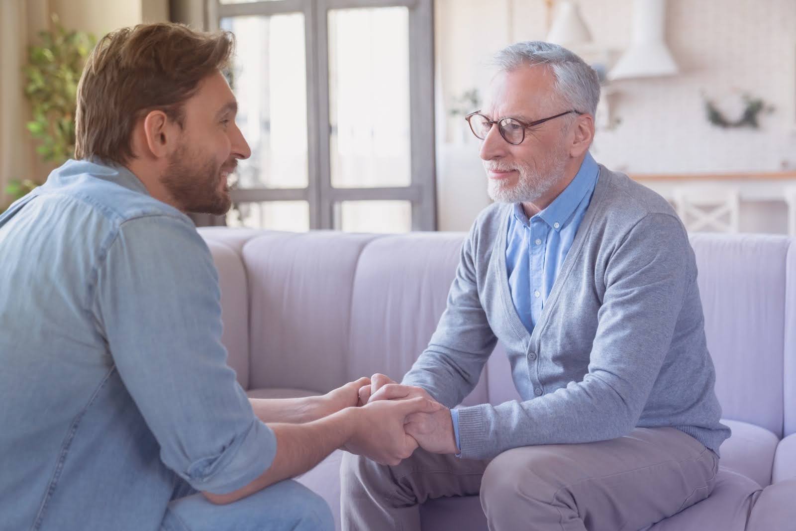 Young adult son having a conversation and supporting his elderly father at home.