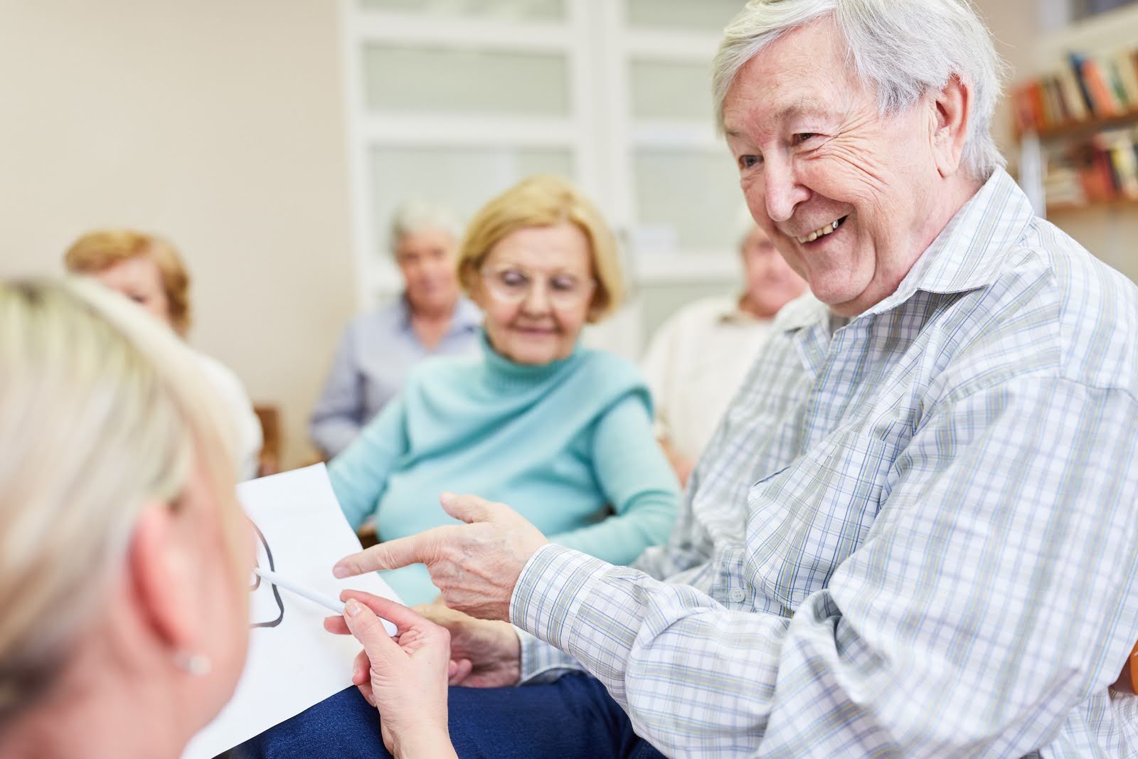 Senior man smiling and pointing at drawing with a caregiver, and other seniors watching in the background with a smile
