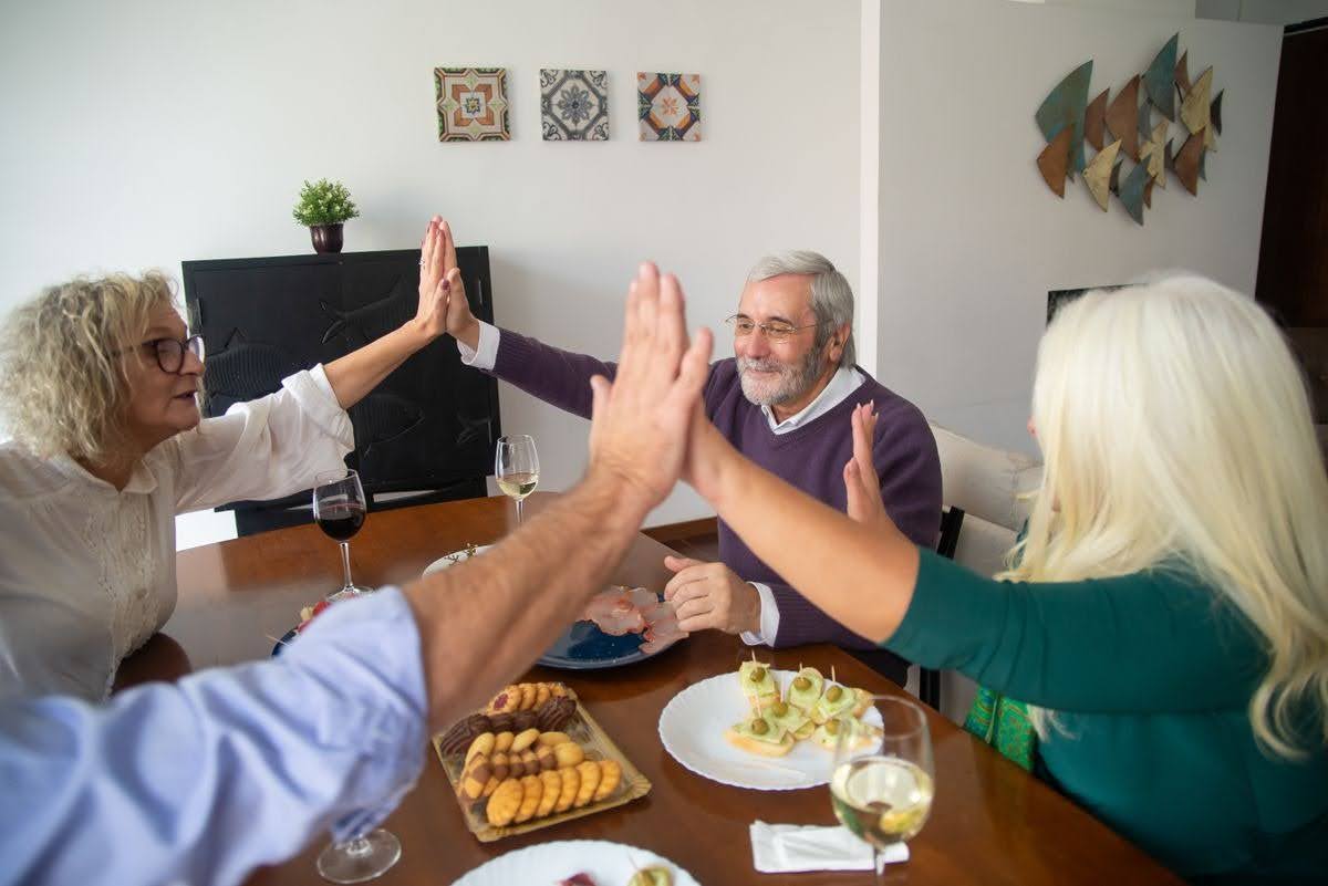 Four older adults high-five across a dining table set with wine glasses, cookies, and appetizers, smiling during a meal at home.