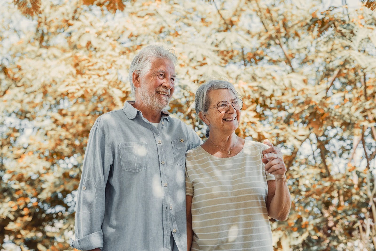 Two smiling adults walk arm in arm through a sunlit autumn park, sharing a warm moment of companionship, happiness, and a relaxed retirement lifestyle outdoors.