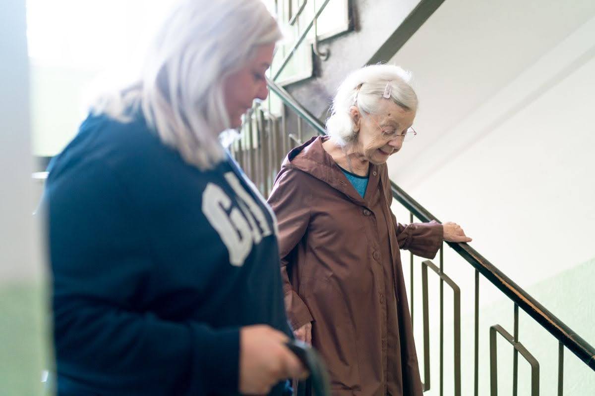 An older adult in a brown coat grips a metal stair railing while a younger adult walks beside them in a stairwell.